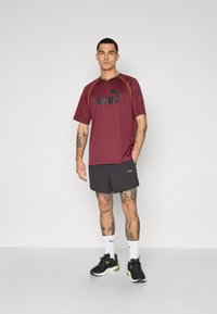 Bordeaux sportt-shirt met zwart Puma-logo, gele accenten. Gepaarde met zwarte shorts en zwarte hardloopschoenen, met een groene detail.