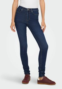 Cienkie dżinsy w ciemnoniebieskim denimie, z wysokim stanem, kieszeniami z przodu i kontrastowymi szwami. Połączone z bordowymi trampkami.