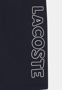 Marineblaue Baumwoll-T-Shirt mit einem großen weißen "LACOSTE"-Logo, das vertikal an der linken Seite aufgedruckt ist. Sauberes, einfaches Design, keine Muster.