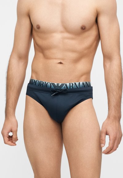 Μυώδης άνδρας που φοράει μπλε σκούρα εσώρουχα Emporio Armani με λογότυπο στη μέση και κορδόνι, στέκεται μπροστά σε απλό ανοιχτό φόντο.
