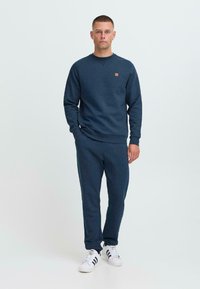 Hombre de pie con un chándal azul marino a juego y zapatillas blancas, manos relajadas, fondo neutro.