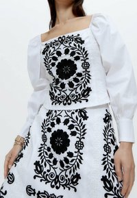 Hvid bluse og nederdel sæt med sort floralt broderi. Blusen har en firkantet udskæring og pufærmer; nederdelen har et matchende design.