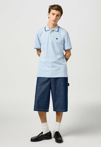 Ljusblå pikétröja med svarta sömnadsdetaljer, matchad med lösa, mörka denimshorts. Modellen bär svarta blanka skor och vita strumpor.