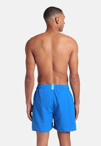 Uomo con capelli corti di spalle, che indossa pantaloni da bagno blu brillante con cintura elastica, in piedi davanti a uno sfondo bianco uniforme.