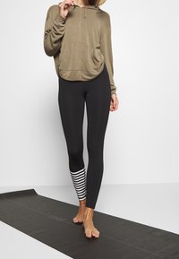 En khakifärgad huvtröja med en framficka, som är kombinerad med svarta leggings med ett randigt mönster på ena vristen. Ståendes på en yogamatta.