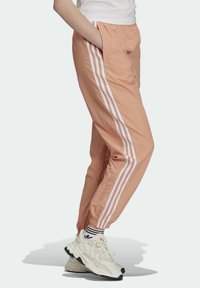 Kalhoty na běhání v broskvové barvě mají bílé diagonální pruhy po stranách, úzký střih, elastický pas a postranní kapsy.