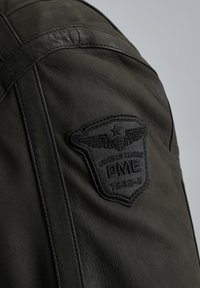 Giacca di pelle verde scuro con superficie texture e un rilevante patch ricamato nero con ali e il testo "PME 1649-A."
