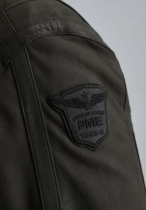 Chaqueta de cuero verde oscuro con una superficie texturizada y un prominente parche negro bordado con alas y el texto "PME 1649-A."