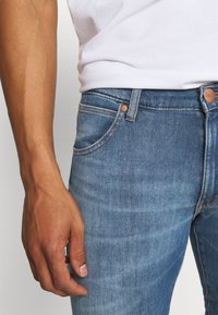 Blue denim jeans with a slim fit, featuring contrast stitching, front pockets, and a small copper-button closure. Textured fabric detail visible.