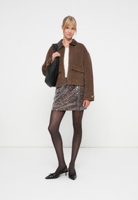 Bruine corduroy jas met een donkere kraag, gecombineerd met een textuurrijk mini-jurkje met pailletjes in grijs- en bruintinten. Zwarte slingback hakken en handtas.