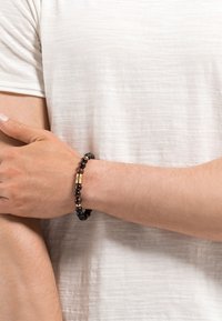 Bracciale di perline nere e marroni con accenti dorati, caratterizzato da una combinazione di texture opache e lucide, indossato su un polso nudo contro una camicia chiara.