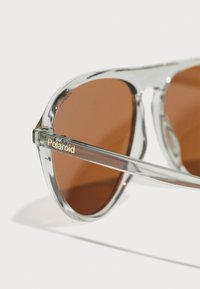 Ochelari de soare cu montură transparentă și lentile maro. Logo-ul "Polaroid" este afișat în aur pe bratul ochelarilor. Prezintă un design clasic de tip aviator.