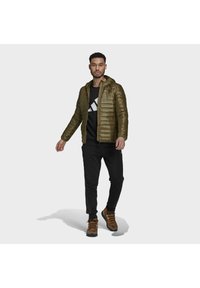 Veste légère matelassée vert olive avec capuche, fermeture éclair à l'avant et couture horizontale. Portée sur une chemise noire et un pantalon noir avec des baskets.