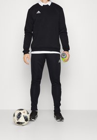 Person i svart Adidas-träningsoverall som håller en grön vattenflaska, stående bredvid en svart-vit Adidas fotboll på ett vitt golv.
