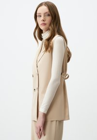 Gilet senza maniche beige con design doppiopetto e bottoni dorati, indossato su una blusa aderente a maniche lunghe bianca. Tessuto dalla texture liscia.