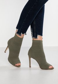 Bottines ouvertes vert olive avec un dessus en tricot texturé, un talon effilé et une hauteur à la cheville. Associées à un jean skinny en denim foncé.