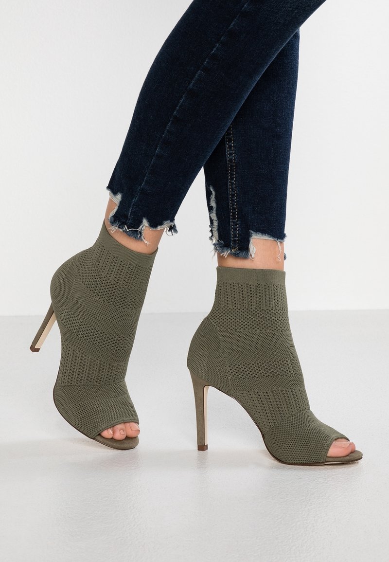 Bottines ouvertes vert olive avec un dessus en tricot texturé, un talon effilé et une hauteur à la cheville. Associées à un jean skinny en denim foncé.