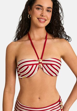 Femme aux longs cheveux bouclés portant un haut de bikini bandeau rayé rouge et blanc avec des boutons dorés et un bas assorti.