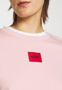 Sudadera rosa con cuello redondo, que presenta un parche bordado rojo con "HUGO". Tela suave y cuello acanalado.
