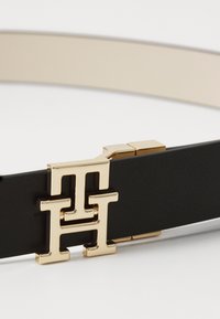 Svart belte med gullfester som har en dobbel "H" logo-spenne og en glatt, beige innerkant. Strukturerte materialer gir detalj.