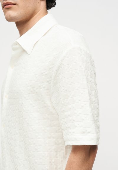 Άνδρας που φοράει λευκό, δομημένο πουκάμισο με κοντά μανίκια και ανοιχτό γιακά, εμφανιζόμενος από τον ώμο μέχρι το μέσο του κορμού σε απλό φόντο.