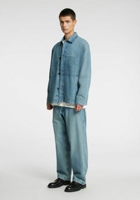 Svetlo modra denim jakna z zapiranjem na gumbe, z dvema žepoma na prsih, nošena preko belega srajčnega kroja. Kombinirana z ohlapnimi svetlo modrimi denim hla�čami in črnimi čevlji.