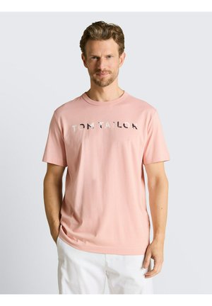 Mężczyzna stoi na jasnym tle, ubrany w różowy T-shirt z krótkim rękawem z napisem „TOM TAILOR” oraz białe spodnie, ręce w kieszeniach.