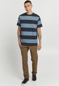 Marineblå og blåstripet kortermet t-skjorte, avslappet passform, brukt med brune bukser og svarte sneakers. Merker av tatoveringer på armene.