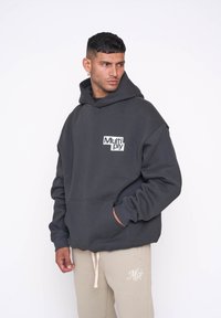 Sudadera con capucha de un gris oscuro, con un bolsillo frontal, que presenta un logotipo blanco de "Multiply" en el lado izquierdo. Tejido suave y grueso, corte relajado.