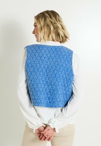 Chaleco acolchado azul con un patrón de diamantes, diseño corto, con bordes redondeados y una tela ligera. Lleva encima una blusa blanca.