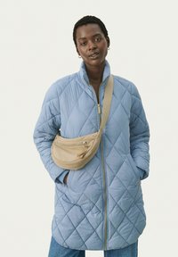 Cappotto trapuntato azzurro chiaro con colletto alto e zip, abbinato a una borsa a tracolla beige. Presenta tasche laterali e una vestibilità comoda.