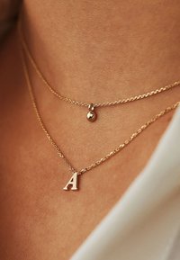 Gelaagde gouden kettingen met een gestructureerde schakel, waarvan de ene een metalen balhanger heeft en de andere een gedurfde, hoofdletter "A" bedel.