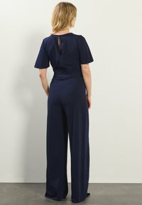 Jumpsuit blu navy con maniche corte, gambe ampie e dettaglio a occhiello sulla schiena. Realizzato in un tessuto liscio con vita aderente.