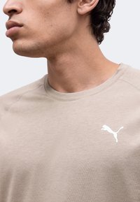 Νέος άνδρας με ανοιχτό μπεζ μπλουζάκι Puma με λευκό λογότυπο Puma στο στήθος, εμφανιζόμενος από τον λαιμό έως το πάνω μέρος του κορμού.
