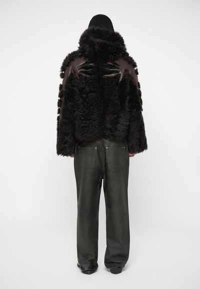 Melnas aitas kažokādas mētelis ar augstu apkakli un ādas akcentiem, saderēts ar tumši zaļām ādas biksēm. Mētelis ir ar strukturētu, pūkainu ārpusi.