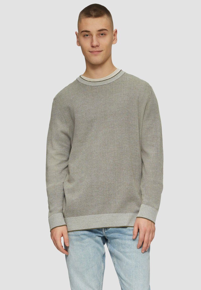 Szary sweter z dzianiny o teksturowym splocie, okrągłym dekolcie oraz kontrastowymi ściągaczami na mankietach i u dołu. Nosi się go na jasnoniebieskich dżinsach.