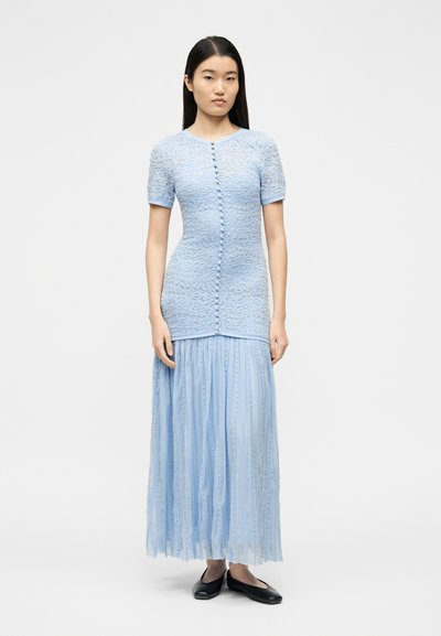 Γυναίκα που φοράει ανοιχτό μπλε φόρεμα από δαντέλα με κοντά μανίκια, κουμπιά στο μπροστινό μέρος και μακριά πλισέ φούστα, συνδυασμένο με μαύρα φλατ παπούτσια, στέκεται σε λευκό φόντο.