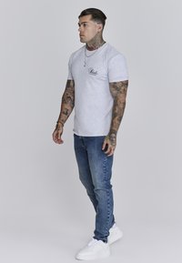 Pilkas medvilninis marškinėlis su juodu logotipu, dėvimas su mėlynomis džinsinėmis kelnėmis ir baltomis sportbatėmis. Modelis turi tatuiruotes ir styled plaukus.