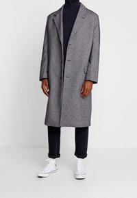 Grå hundtandsmönstrad kappa med slag, sex knappar och sidofickor. Bärs över en marinblå polotröja och tapered mörka byxor med vita sneakers.