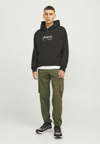 Giovane uomo con felpa nera con logo bianco, pantaloni cargo verde oliva e sneakers nere in piedi contro uno sfondo chiaro.