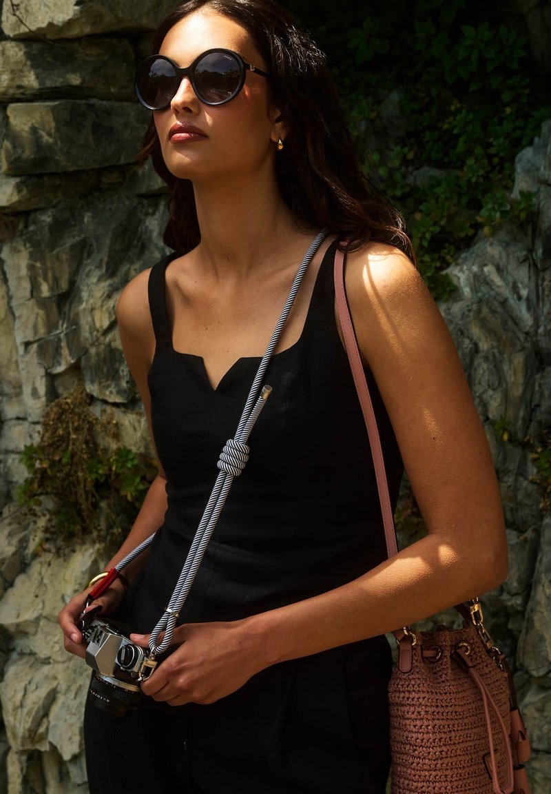 Femme portant une robe noire et de grandes lunettes de soleil rondes, tenant un appareil photo avec une sangle nouée, debout devant un arrière-plan rocheux en plein air.