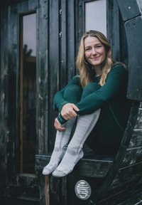 Calze grigie con motivi texturizzati poggiano su una superficie di legno. Una persona indossa un outfit verde scuro, con capelli lunghi, su uno sfondo di legno nero.