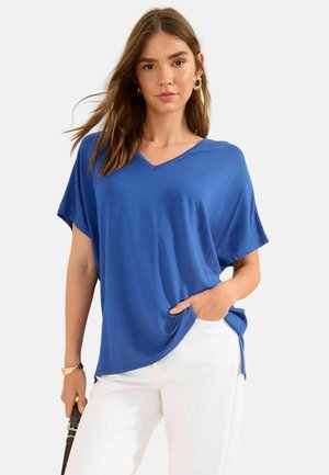 Naine, kes kannab sinist V-kaelusega lühikeste varrukatega tuba ja valgeid pükse, hoides ühe käega koti rihma ning teisega taskus.