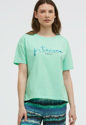 Young woman wearing a mint green T-shirt with blue sequined text and patterned blue-green pants, standing against a plain background.