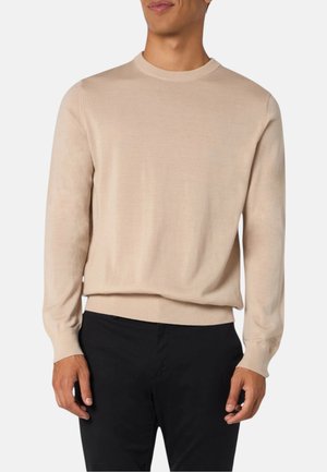 Uomo che indossa un maglione beige chiaro a maniche lunghe e pantaloni neri, in piedi con le braccia rilassate davanti a uno sfondo bianco uniforme.