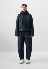 Black waterproof jacket featuring a hood, two front pockets, triangular logo, paired with loose-fitting black pants and white sneakers.