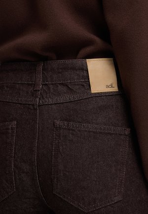 Jeans de color marrón oscuro con una etiqueta de cuero visible etiquetada "adL" en la cintura. Presenta un diseño de corte recto y bolsillos traseros. Tejido texturizado.