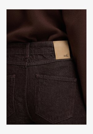 Jeans de color marrón oscuro con una etiqueta de cuero visible etiquetada "adL" en la cintura. Presenta un diseño de corte recto y bolsillos traseros. Tejido texturizado.