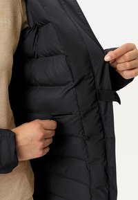 L'interno della giacca nera gonfiabile presenta sezioni trapuntate, una tasca con zip e un flap in velcro per una chiusura sicura. La texture del tessuto è liscia.