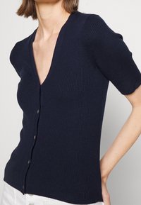 Marinblå ribbstickad cardigan med v-ringning, korta ärmar och knäppning. Har en figurnära design och ett strukturerat stickmönster.